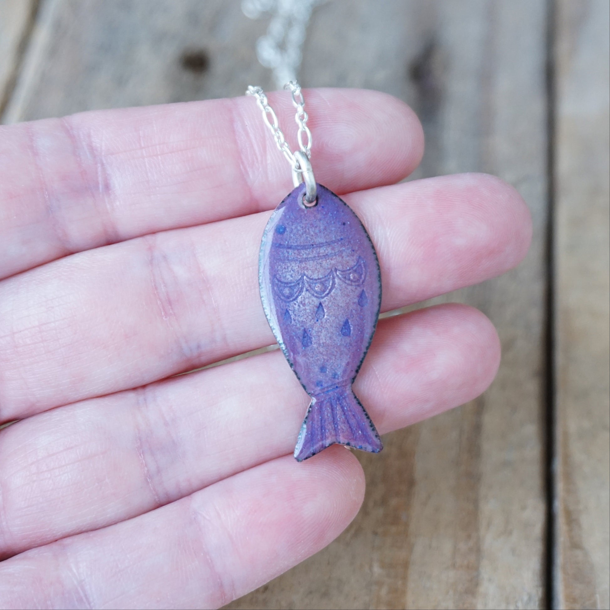 Purple fish-shaped pendant on a chain held in a hand against a wooden background