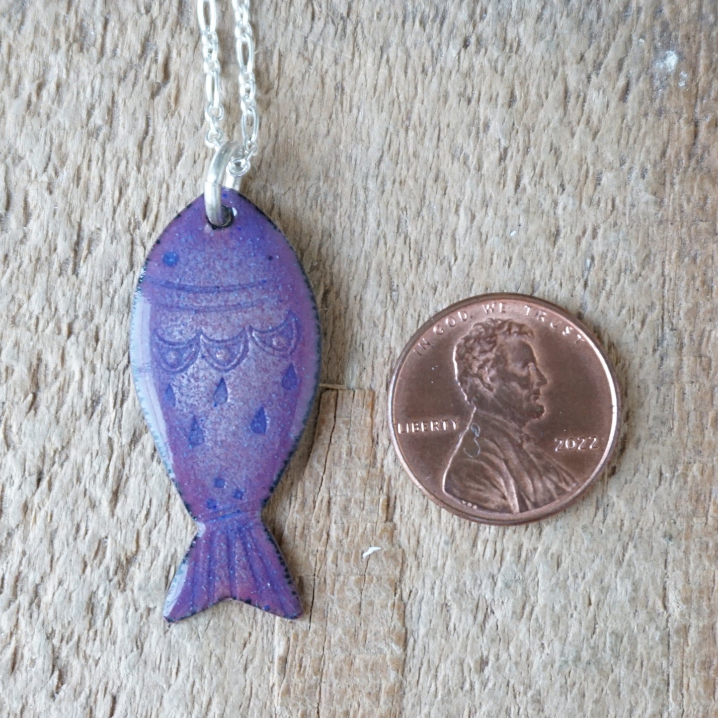 Purple fish-shaped pendant on a chain next to a penny for scale on a wooden surface