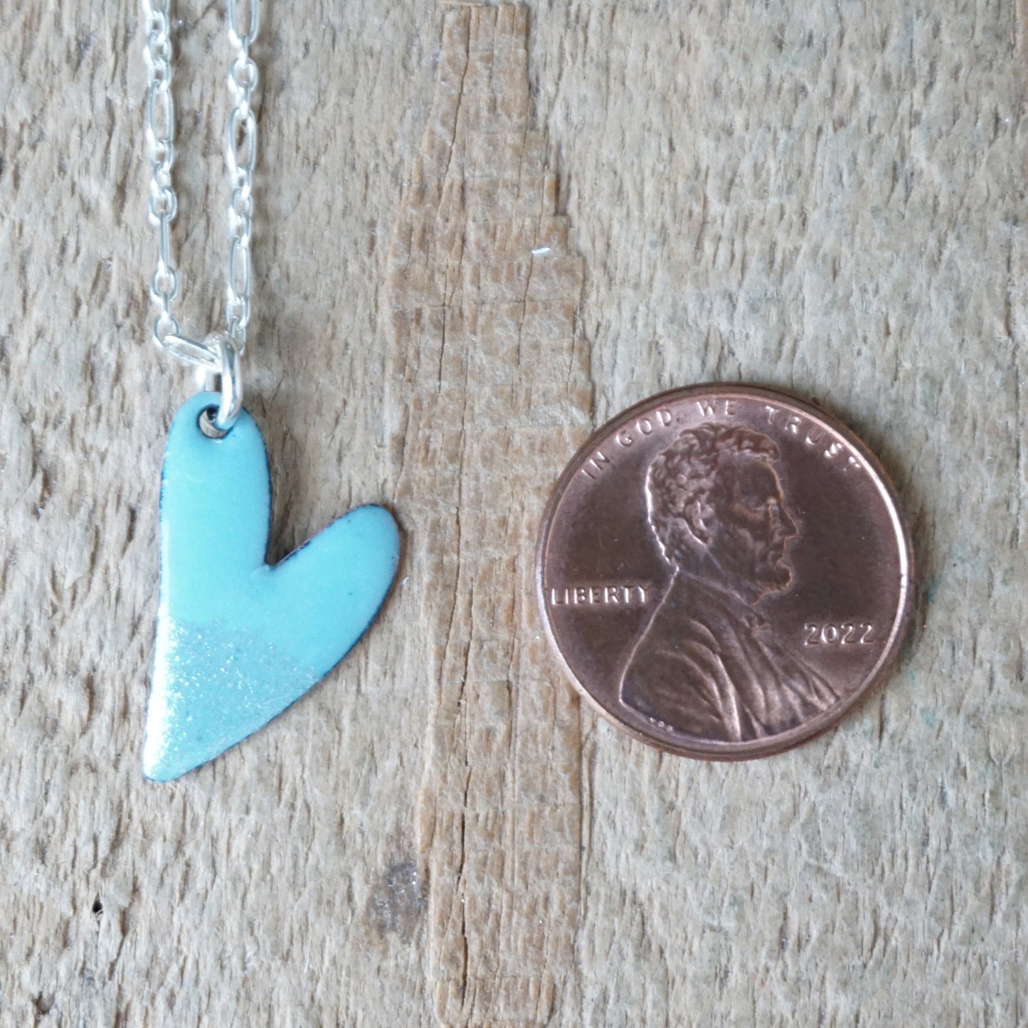 Light blue heart-shaped pendant on a chain next to a penny for size comparison on a wooden surface