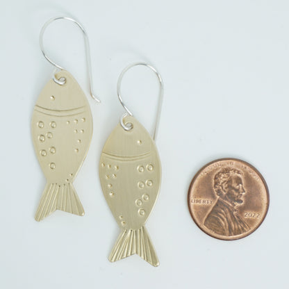 brass fish-shaped earrings next to a penny for scale on a white background