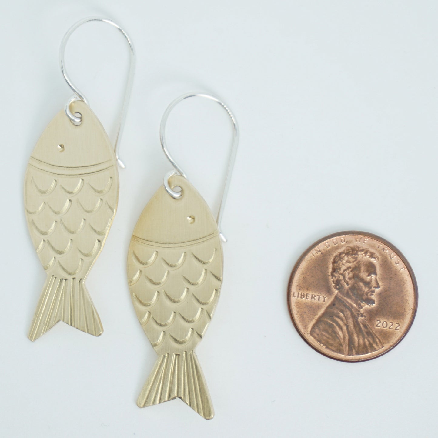 brass fish-shaped earrings next to a penny for scale on a white background