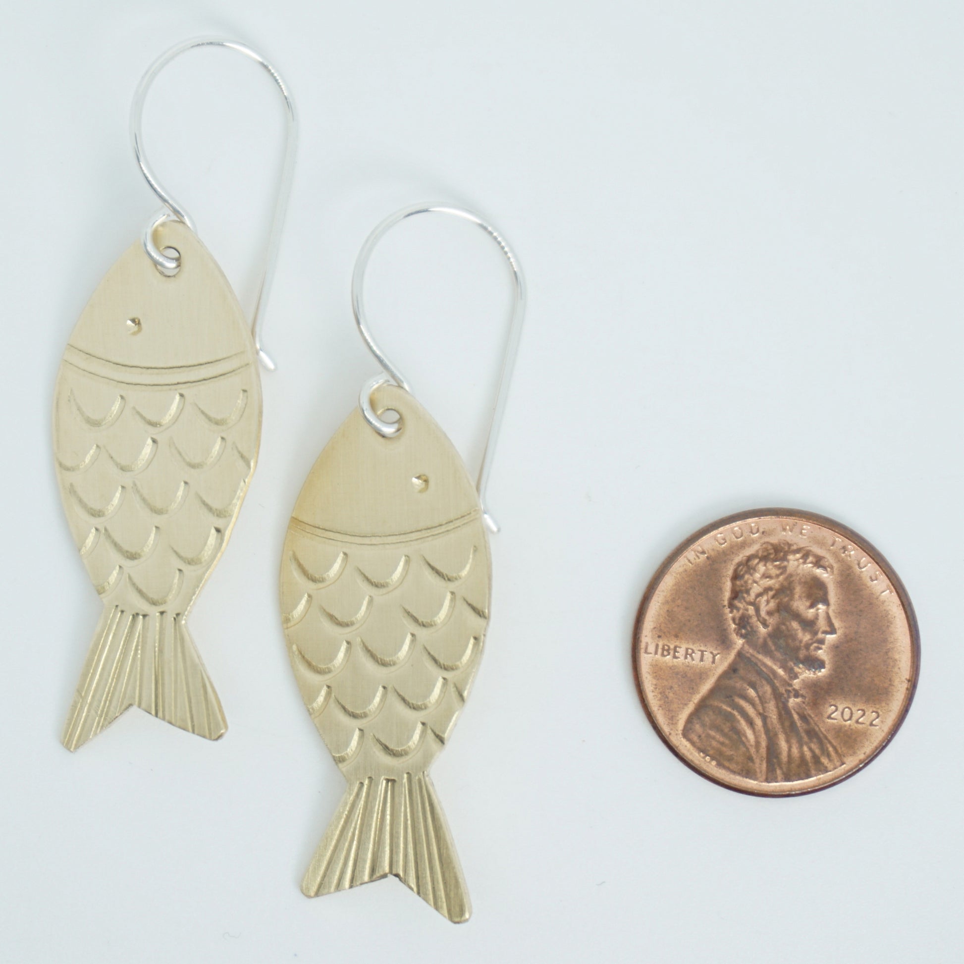 brass fish-shaped earrings next to a penny for scale on a white background