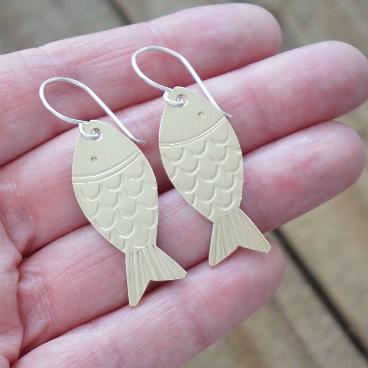 brass fish-shaped earrings held in a hand on a wooden surface