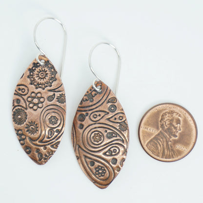 Pair of copper earrings with intricate patterns next to a penny for scale on a white background