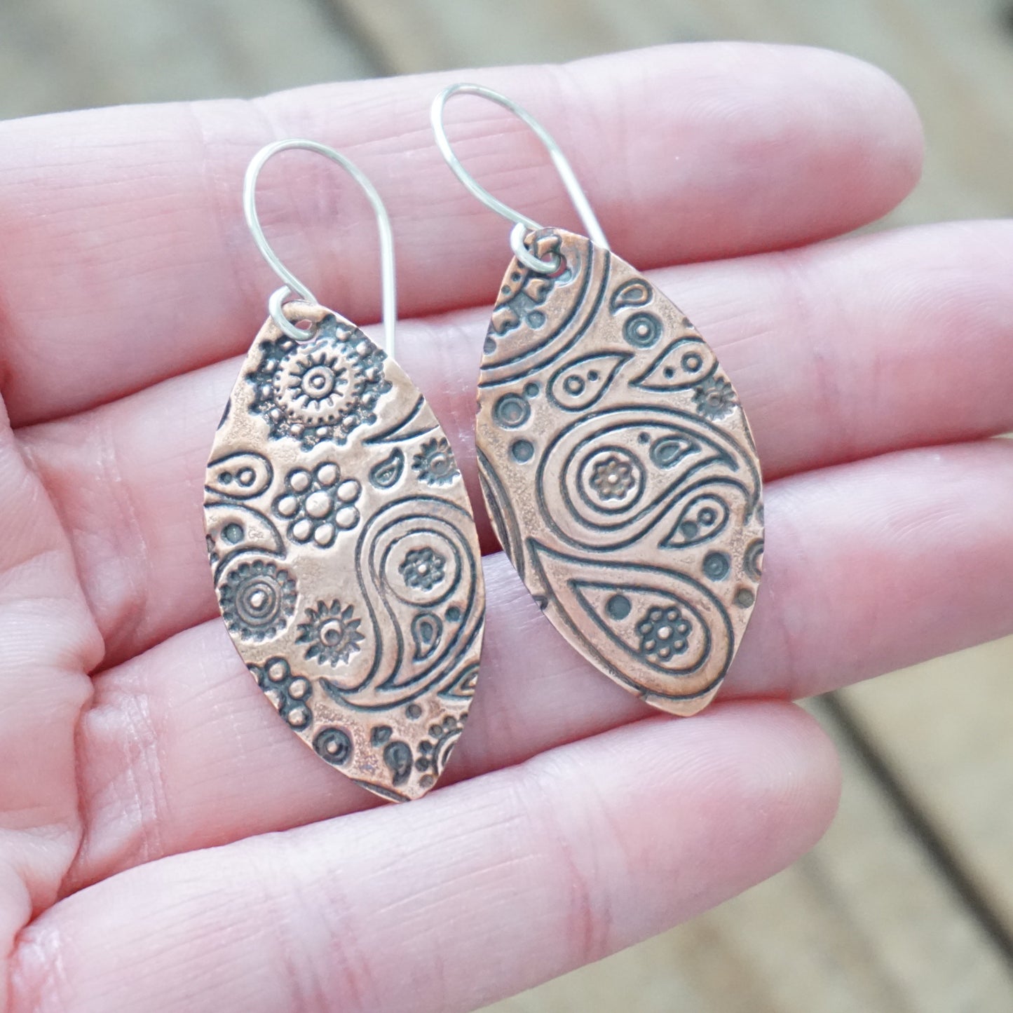 copper paisley patterned earrings held in a hand with a blurred background
