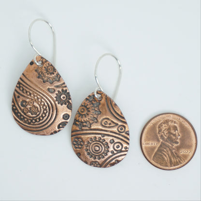 Teardrop-shaped copper earrings with intricate patterns next to a penny on a white background