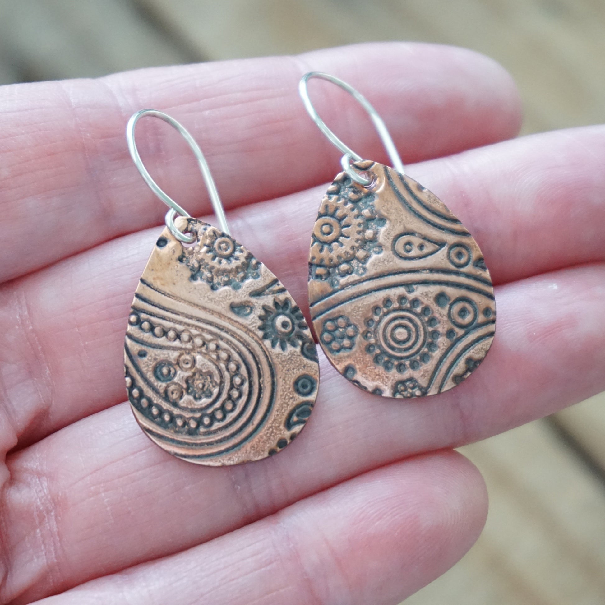 Copper teardrop earrings with intricate patterns held between fingers against a blurred background