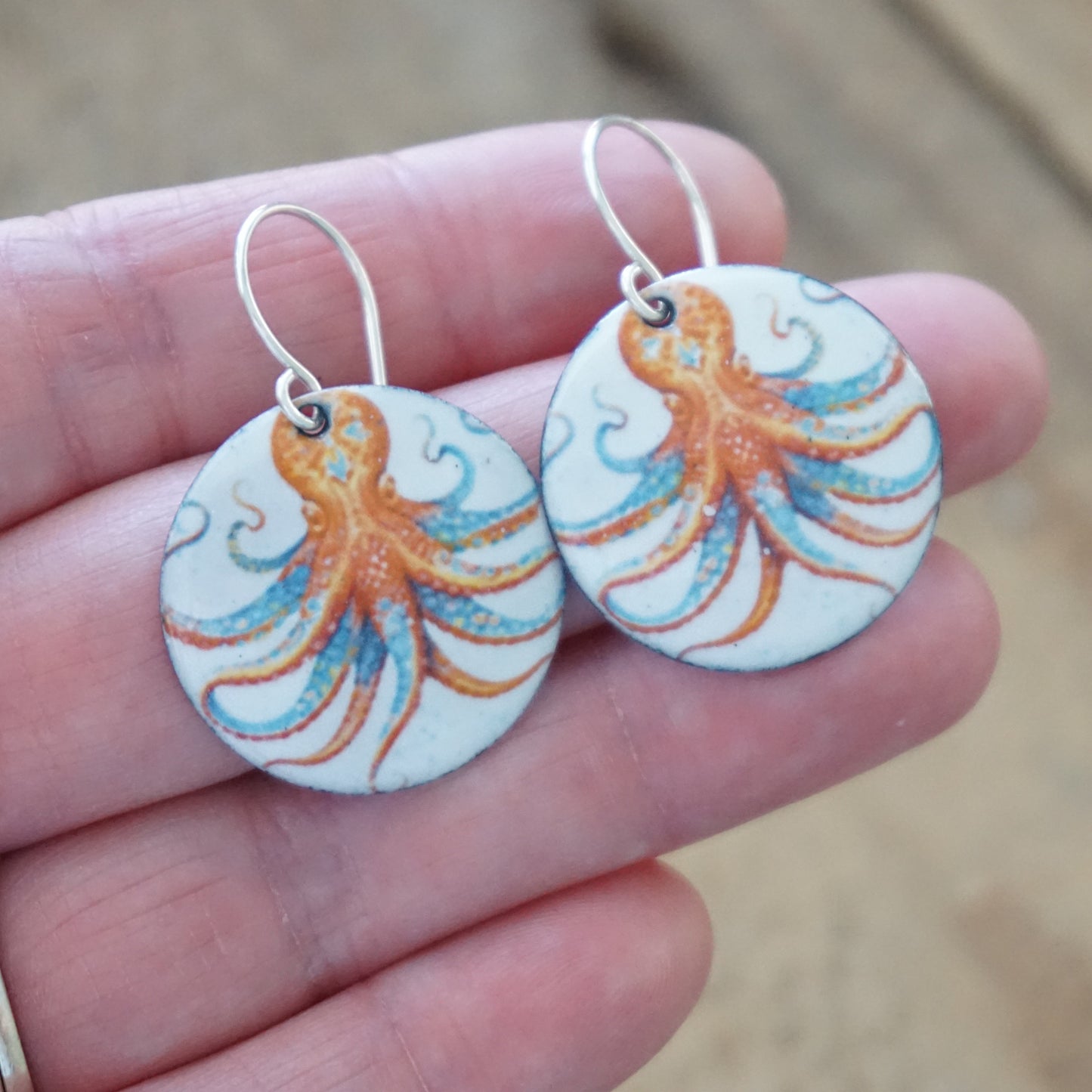Round earrings with octopus design held between fingers against a blurred background