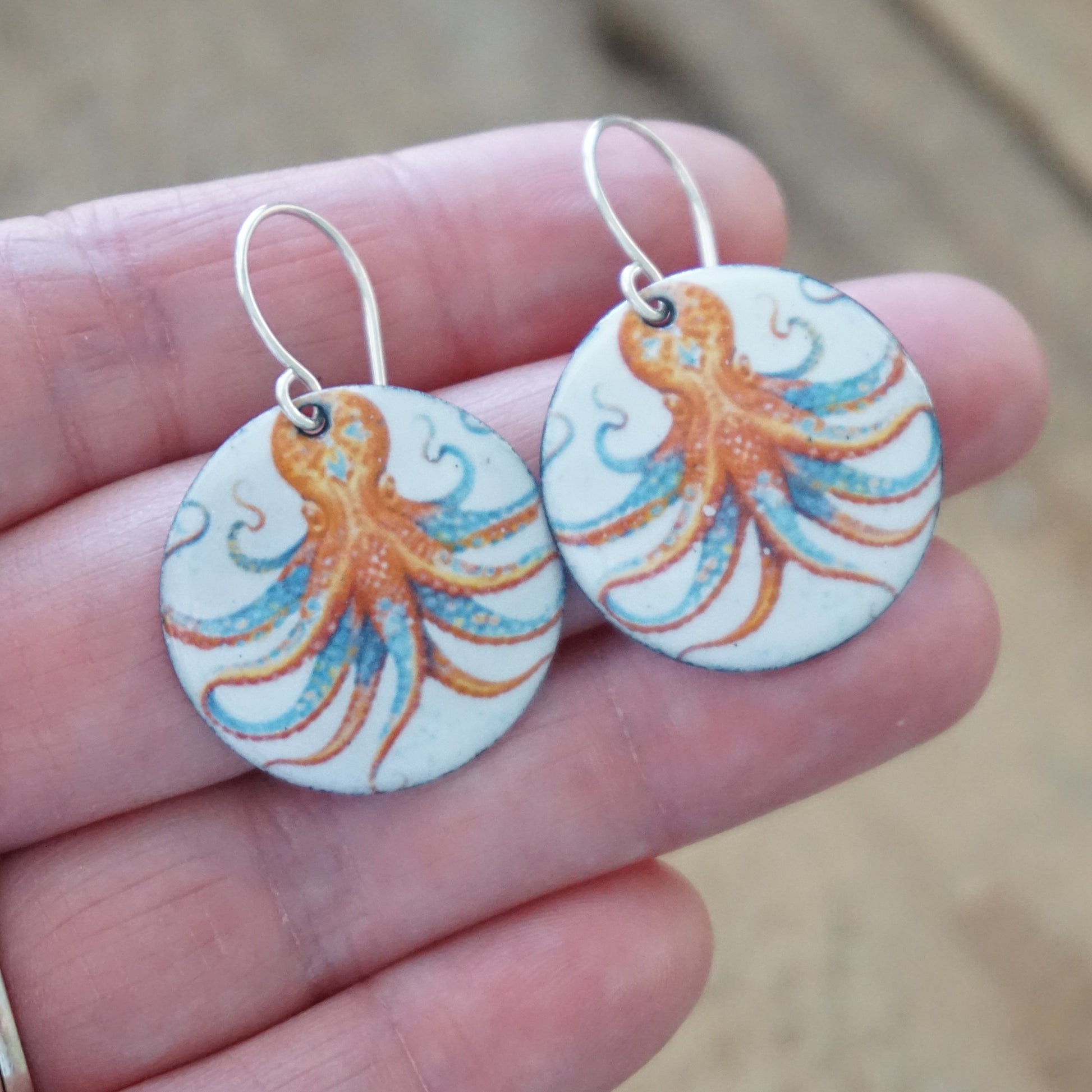 Round earrings with octopus design held between fingers against a blurred background
