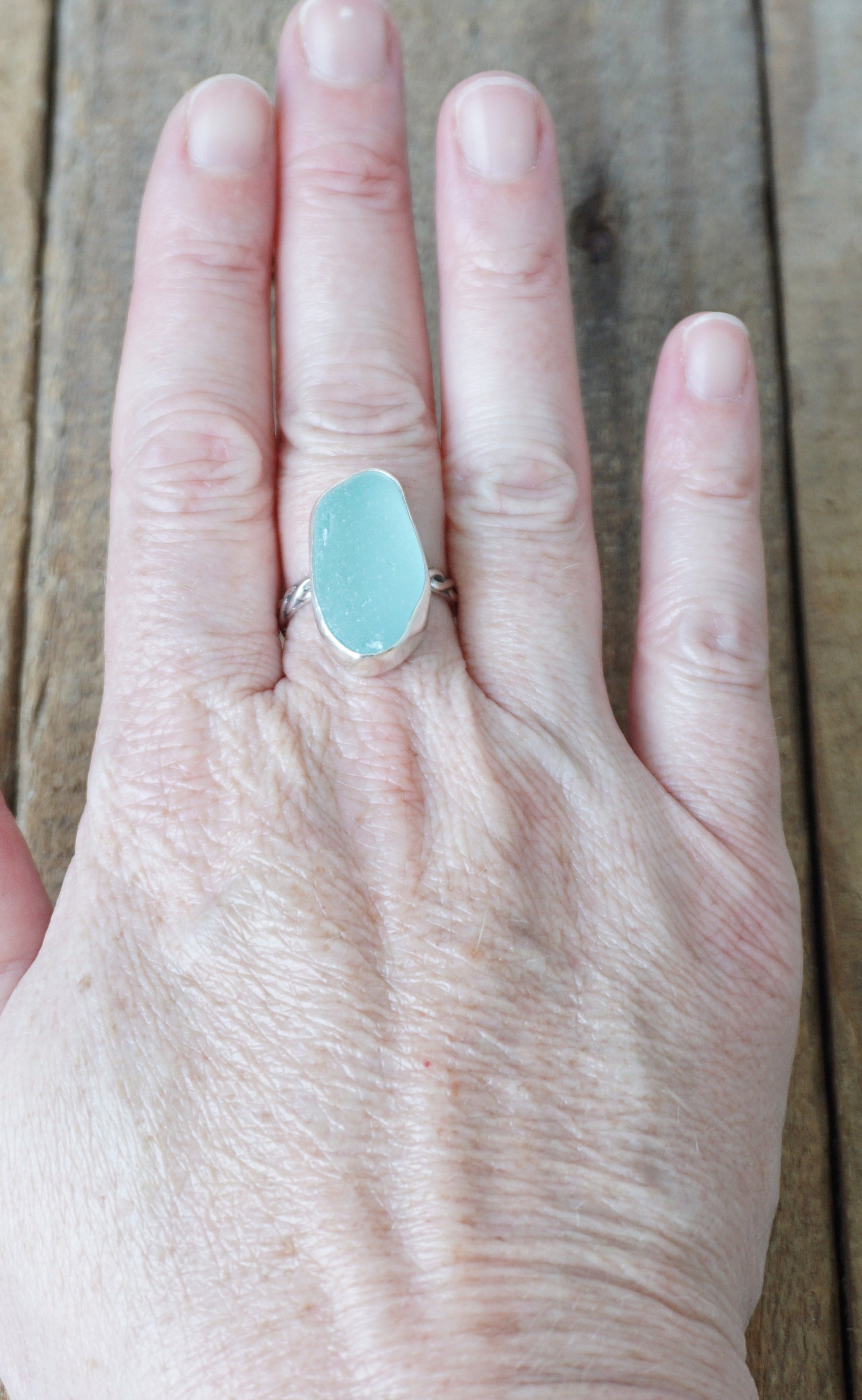 Hand wearing a ring with a light blue sea glass on a wooden surface