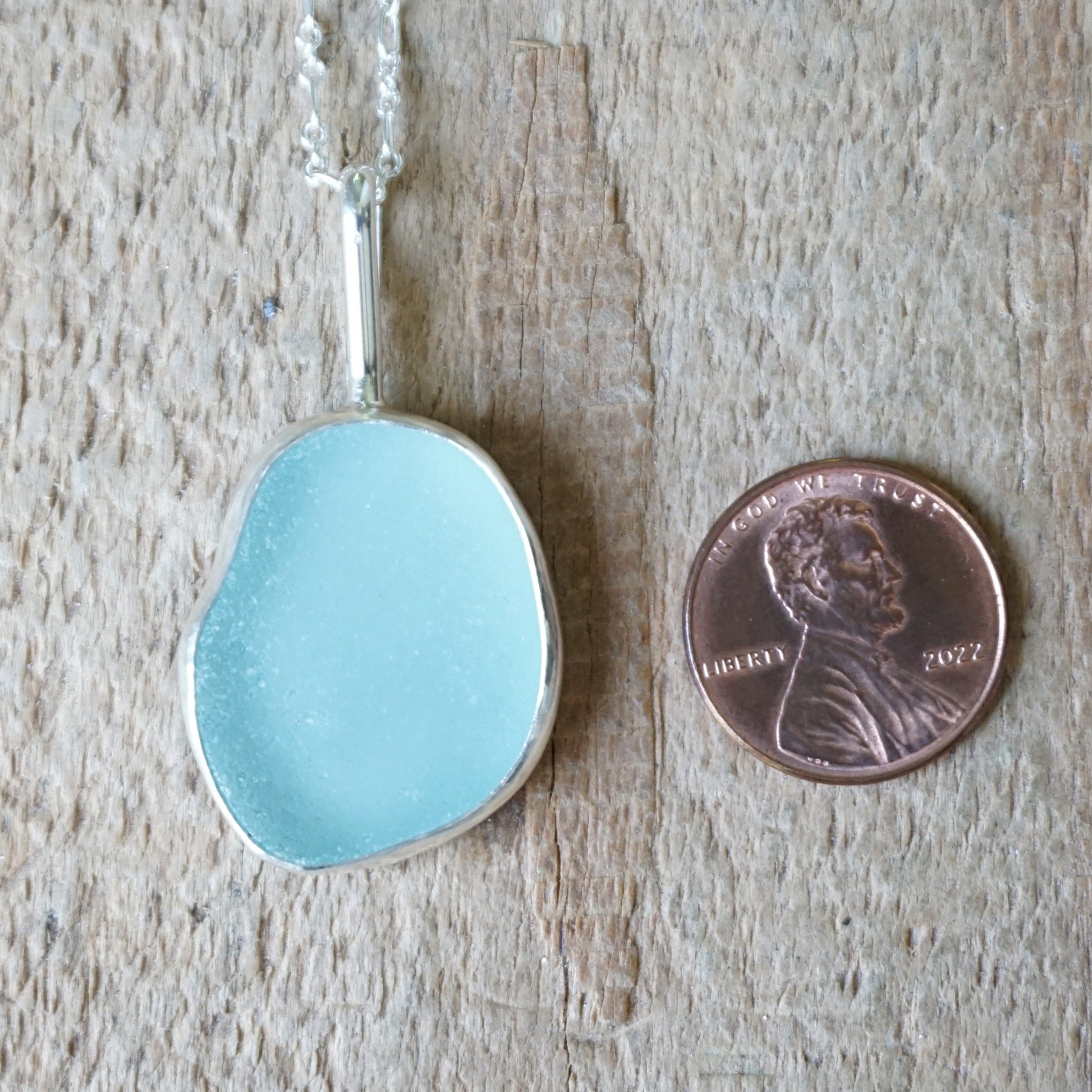light blue sea glass pendant next to a penny for scale on a wooden background