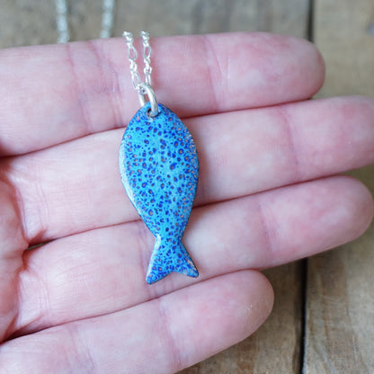 Blue fish-shaped pendant on a chain held in a hand against a wooden background