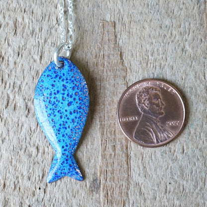 Blue fish-shaped pendant on a chain next to a penny for scale on a wooden surface