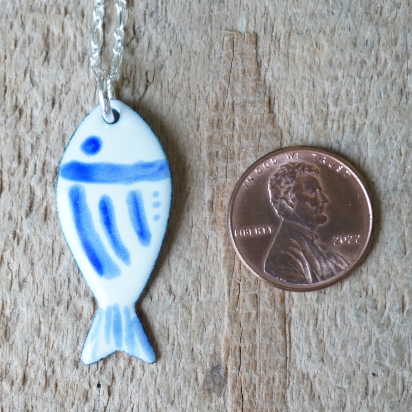 Fish-shaped pendant with blue accents on a chain, next to a penny for scale, on a wooden surface.