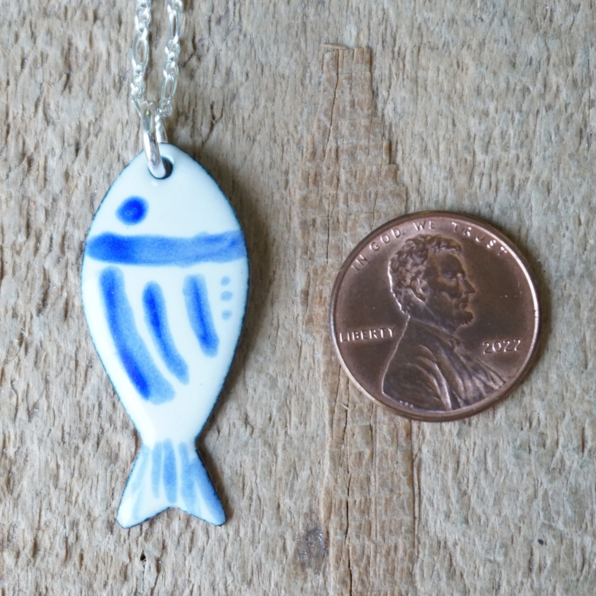 Fish-shaped pendant with blue accents on a chain, next to a penny for scale, on a wooden surface.