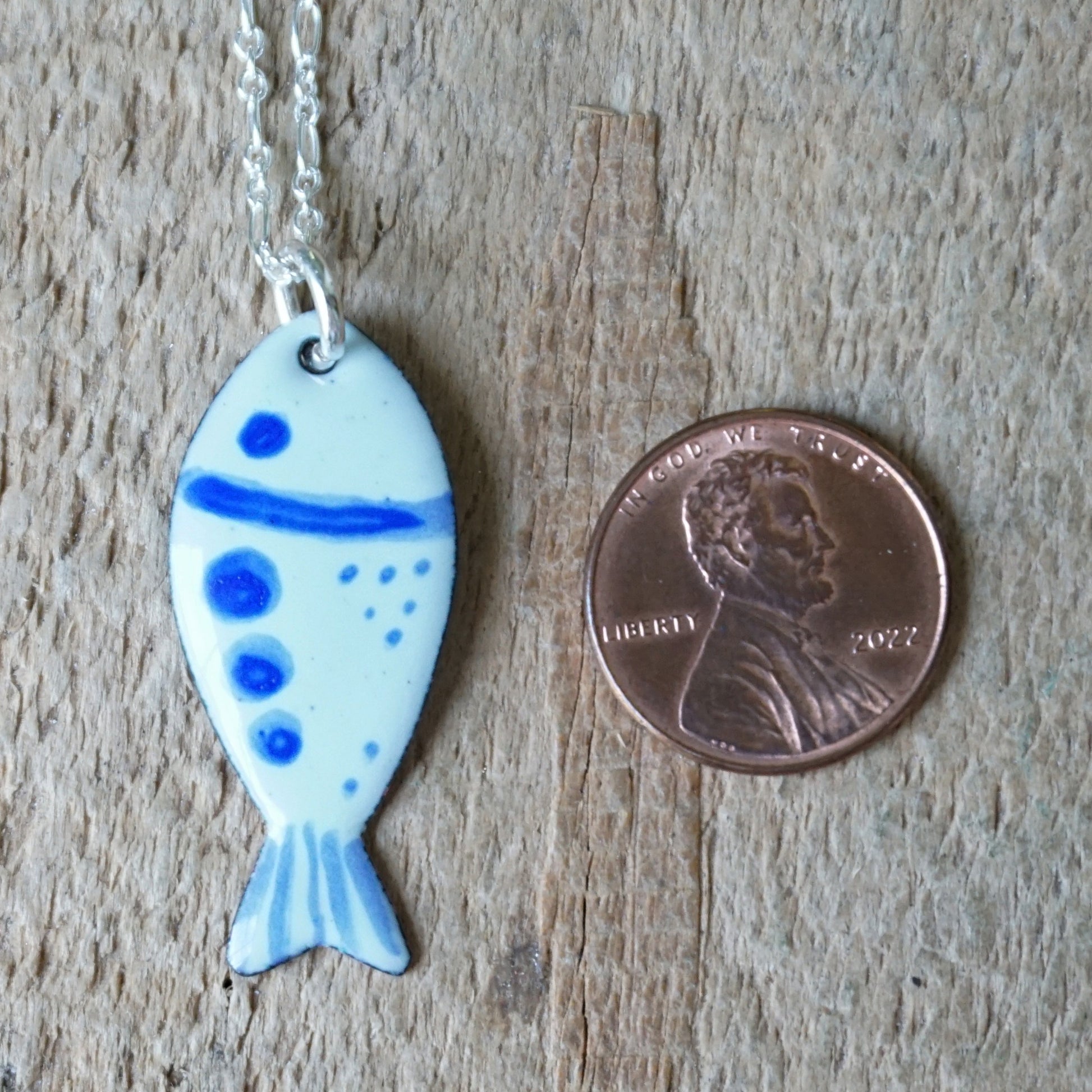 Fish-shaped pendant with blue accents on a chain, next to a penny for scale, on a wooden surface.