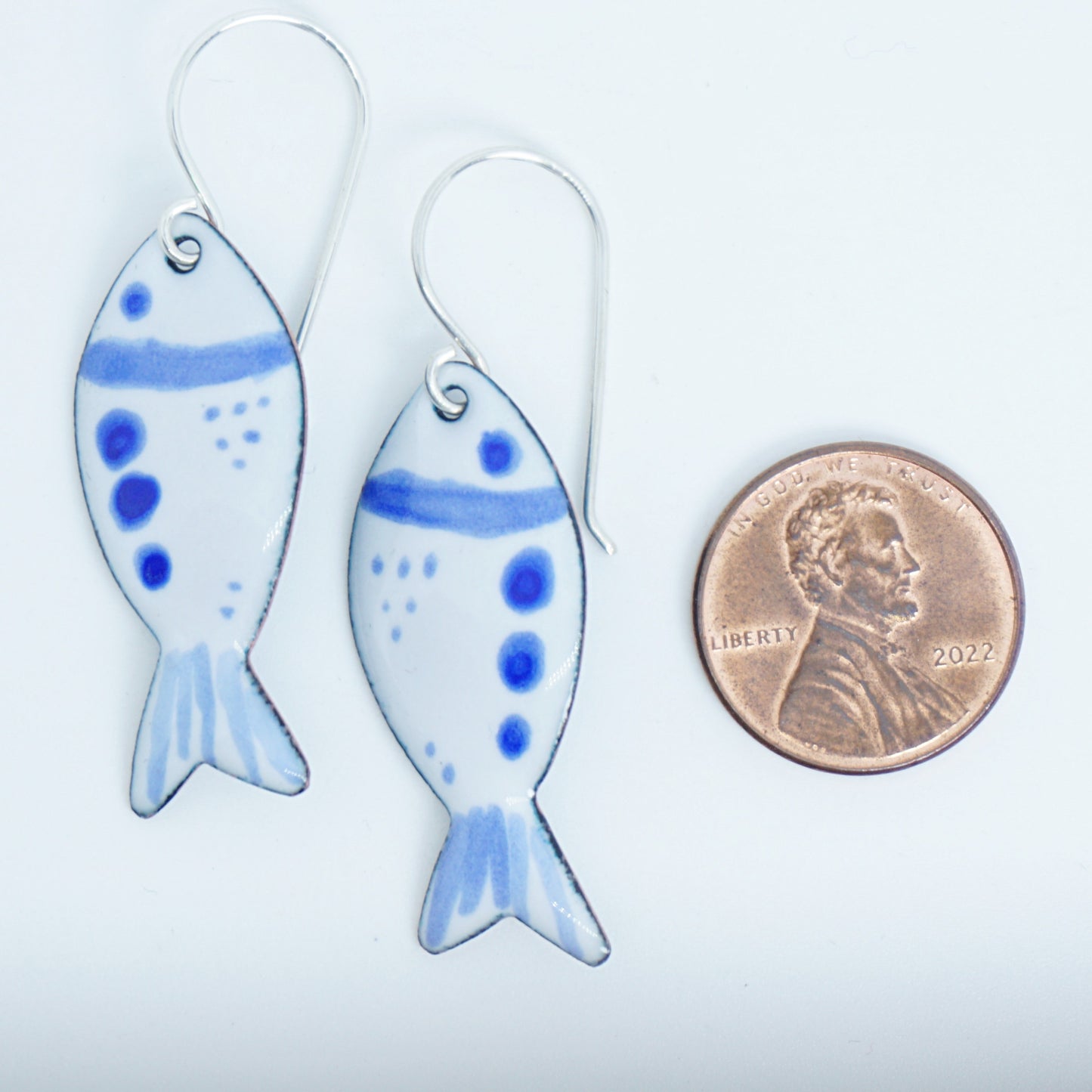 Fish-shaped earrings with blue accents next to a penny for scale on a white background