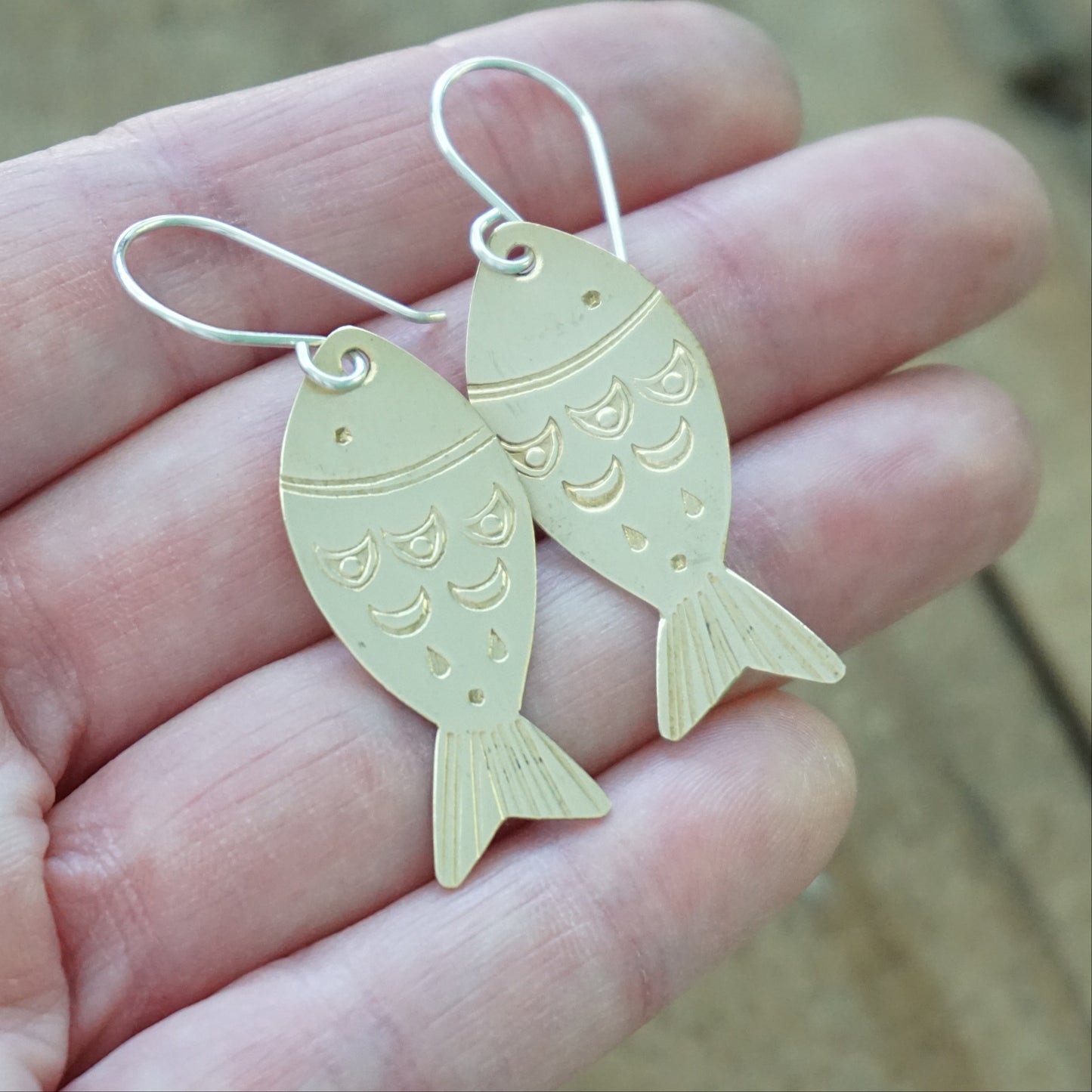 Gold fish-shaped earrings held in a hand with a wooden background