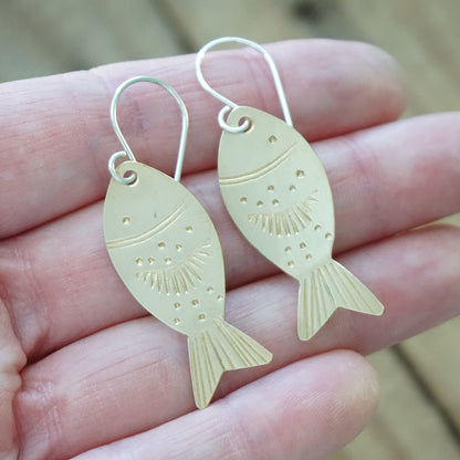 Gold fish-shaped earrings held between fingers against a wooden background