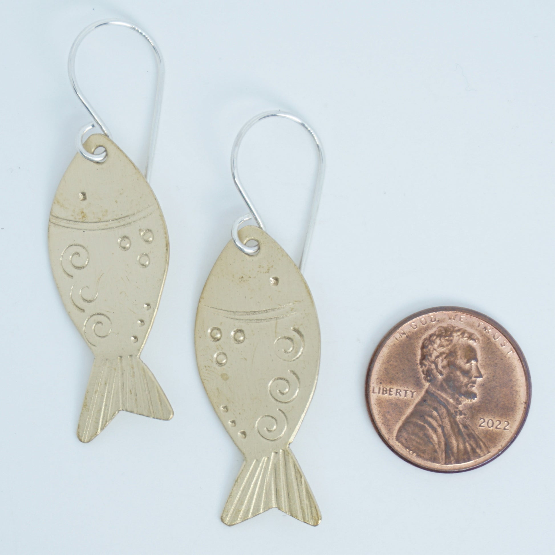 Fish-shaped earrings next to a penny for size comparison on a white background