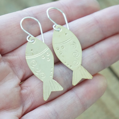 Gold fish-shaped earrings held between fingers against a wooden background